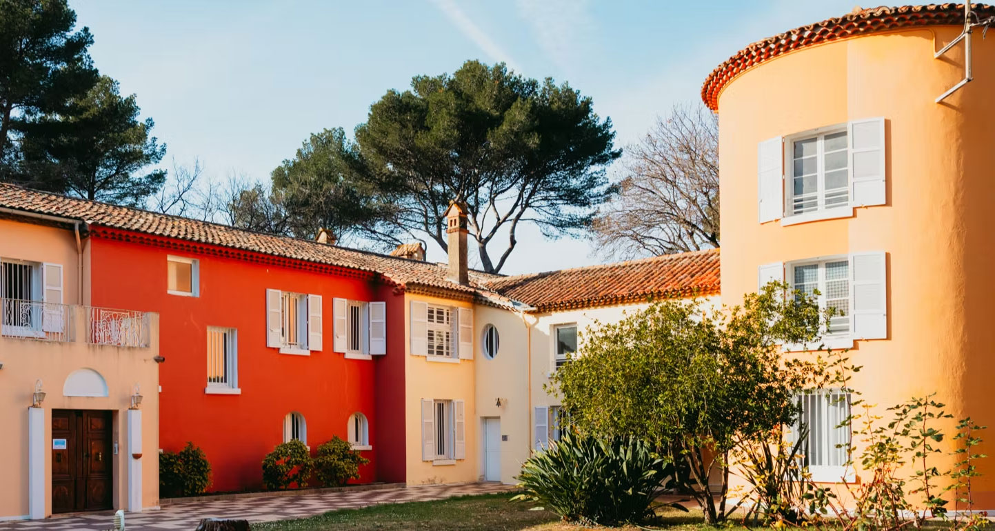 Colorful Mediterranean-style apartment buildings with orange, red, and yellow walls surrounding a courtyard and greenery.
