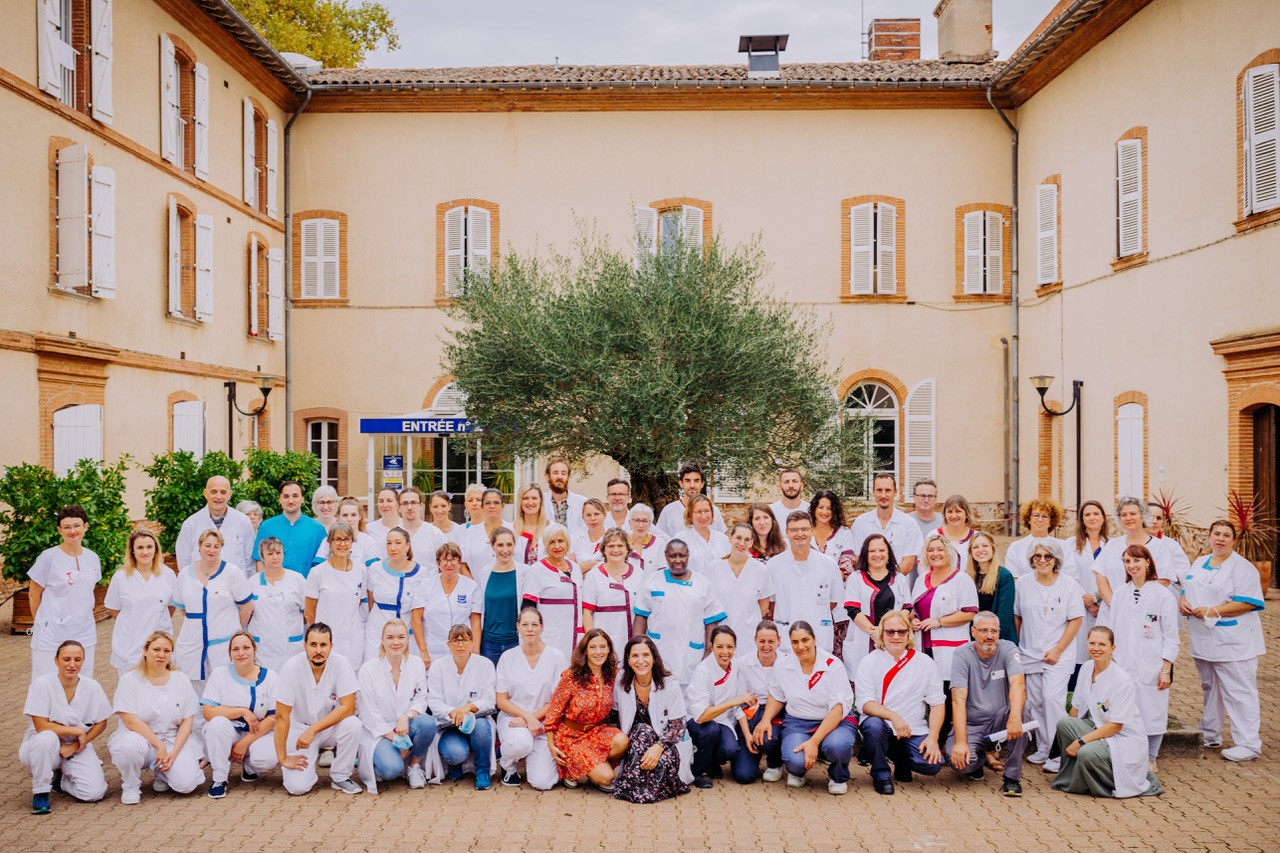 Large group of medical staff in white scrubs posing for a group photo in a sunny courtyard with a central olive tree.
