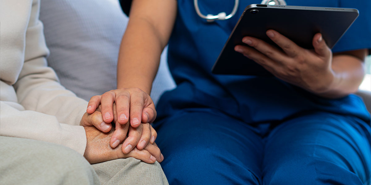 HAD-grand-bordeaux Nurse in blue scrubs with a stethoscope holds a tablet while comforting a patient, their hands clasped together in support.
