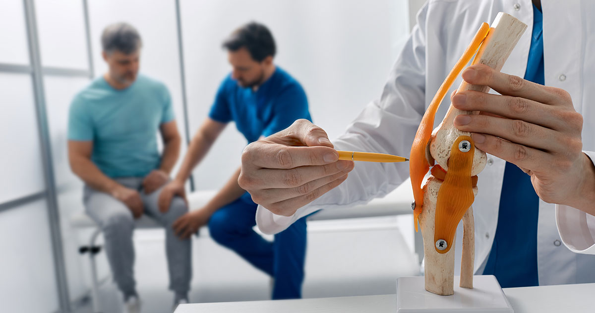 Orthopedic clinician demonstrates knee joint model with orange ligaments to two patients in a clinic.